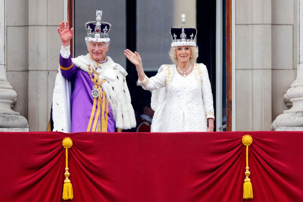 king charles iii and queen camilla stand on a balcony, wave and smile, both wear ornate outfits and crowns