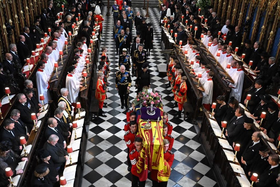 several men carry an adorned coffin as a procession walks behind them, people stand and watch to the sides