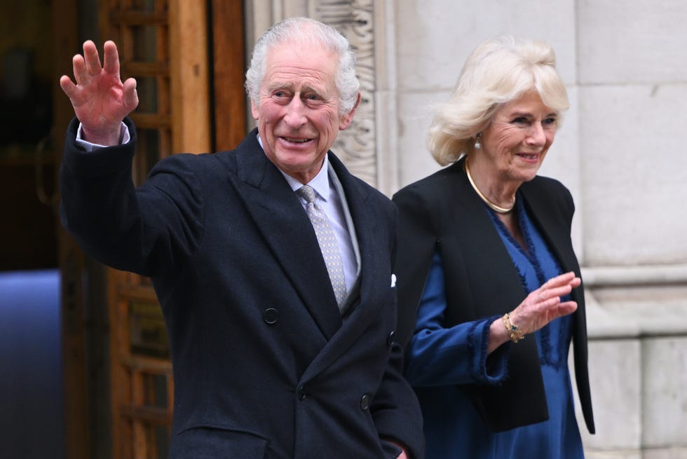 King Charles III and Queen Camilla wave and smiles king charles iii and queen camilla wave and smile after walking out a building, he wears a dark peacoat with a purple collared shirt and silver patterned tie, she wears a black coat over a blue long sleeve dress and golden necklace