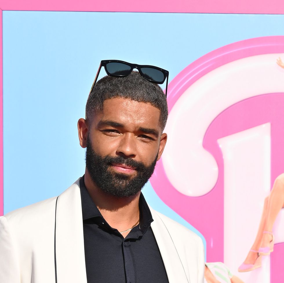 kingsley ben adir, a man with dark hair and a bear, wearing black pants a black shirt and a white suit jacket, on posing for the camera on a pink carpet at the premiere of the barbie movie in 2023