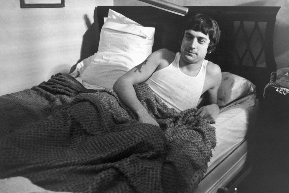 robert de niro in character for mean streets lies in bed on one elbow, he wears a white tank top and has a blanket over his body to his waist