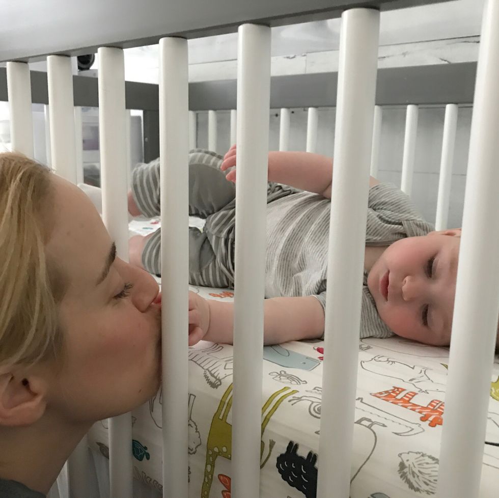 mom kissing a baby's hand through the crib slats, part of a good housekeeping story on how long a baby stays in the crib