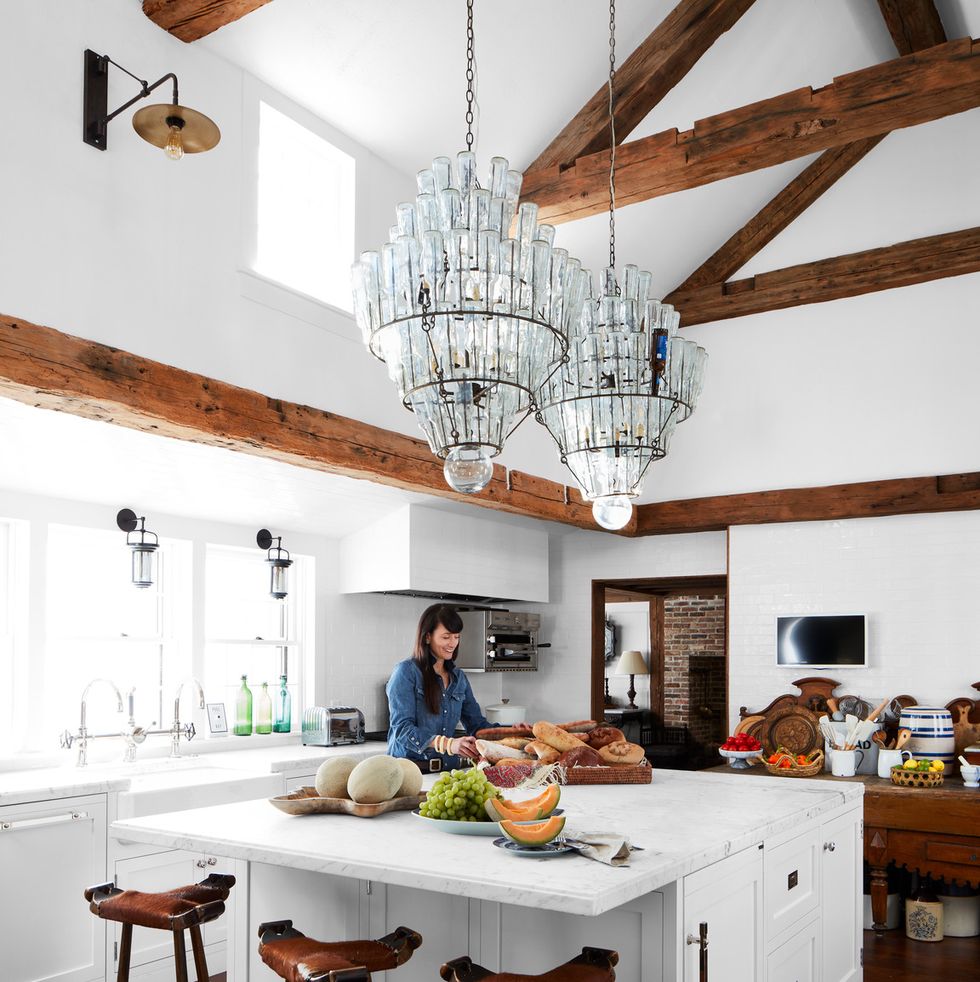 an all white kitchen with wood beams and a large island in the middle where a woman is standing