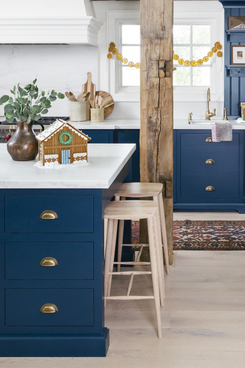 large dark blue kitchen island with drawers, gold toned drawer pulls, white counters, and blond wood stools for seating