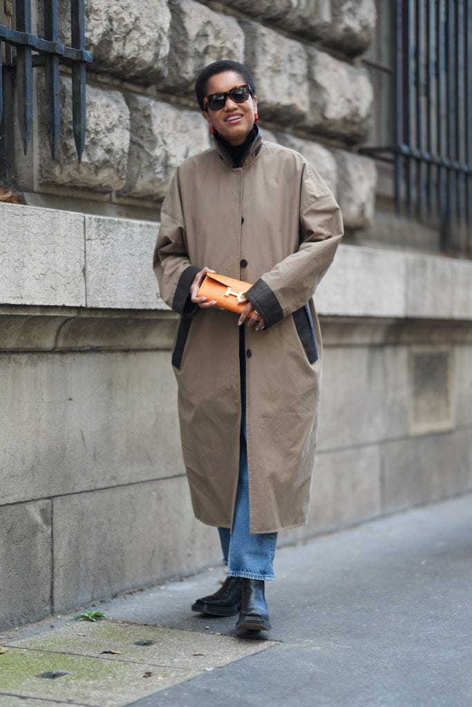 paris, france september 28 tamu mcpherson wears black sunglasses, light brown oversized trench coat, light blue denim jean pants, shiny black heeled booties leather shoes, bright orange textured hermes clutch leather bag, outside hermes, during the paris fashion week springsummer 2025 on september 28, 2024 in paris, france photo by edward berthelotgetty images