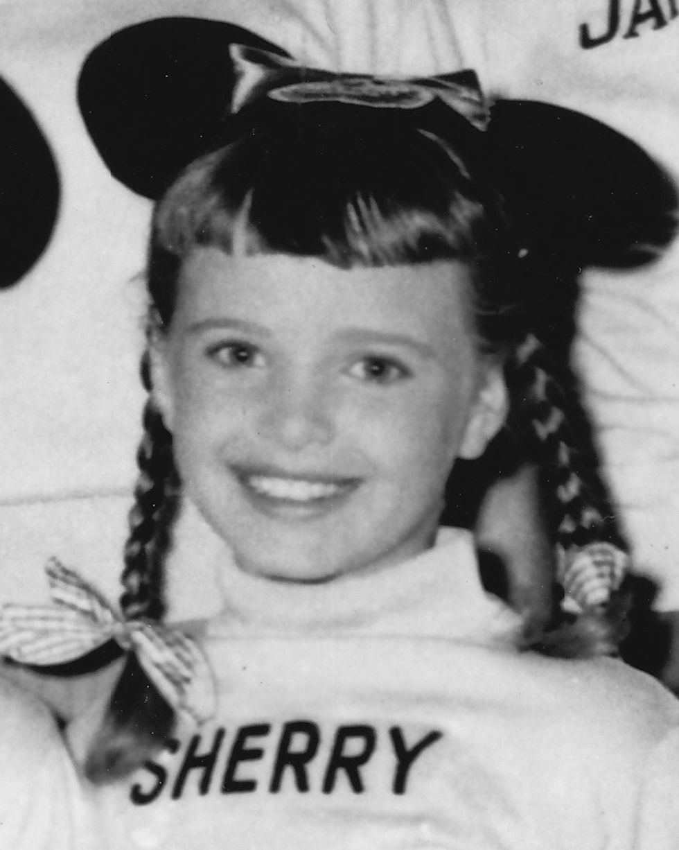 sherry alberoni smiles at the camera she wears a turtleneck with her first name embroidered on it and mickey mouse ears, her hair is in braided pigtails with bows at the ends