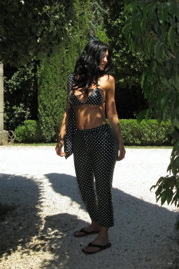 uma pessoa posa em um jardim vestida com um top de biquíni de bolinhas e calças estampadas