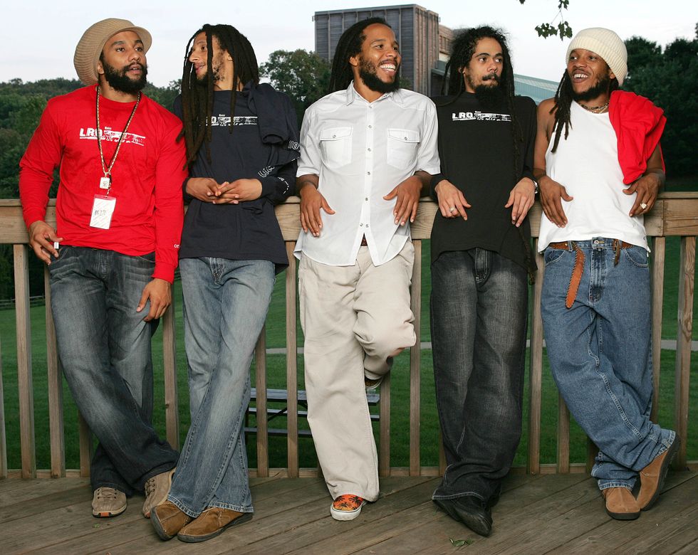 kymani marley, julian marley, ziggy marley, damian marley, and stephen marley lean against a wooden railing and smile while looking in different directions, behind them is a grass field, trees, and tall building