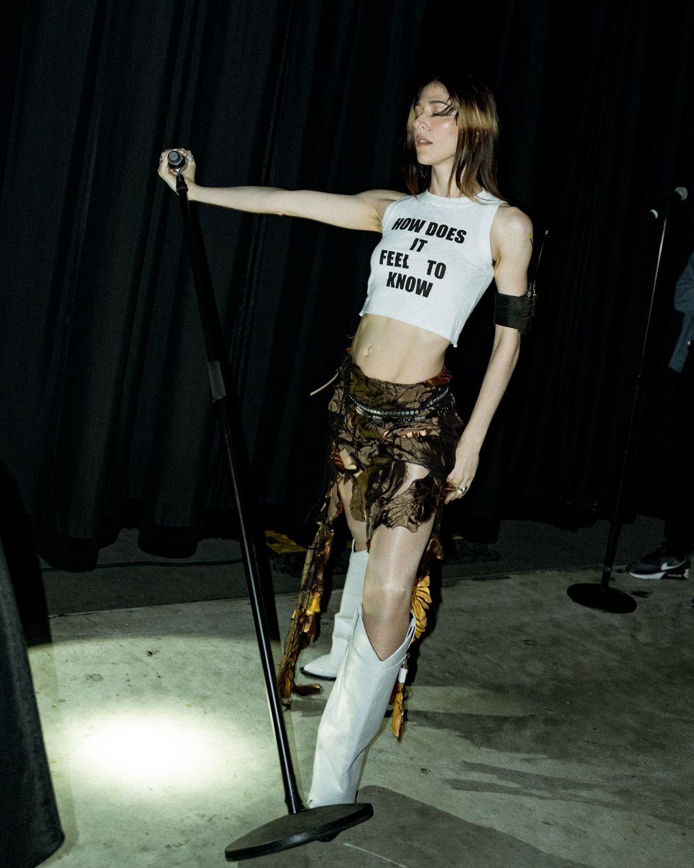 caroline polachek in a crop top that reads 'how does it feel to know,' a skirt with cutouts, and white cowboy boots
