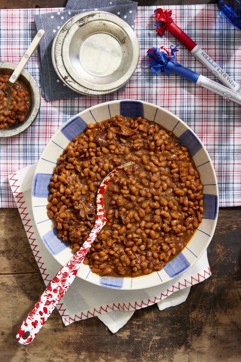 root beer baked beans in a serving bowl with a spoon on a wooden table with a red and blue plaid tablecloth