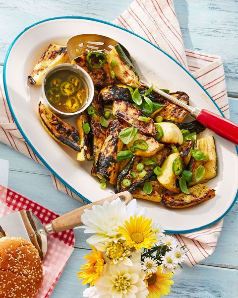 grilled summer squash in a white oval serving dish with a large spoon and a small bowl of lemon scallion dressing