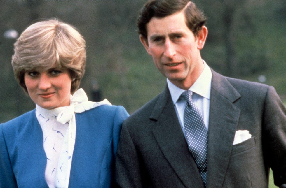 lady diana spencer, wearing a blue dress, and prince charles, wearing a gray suit, white shirt, and blue spotted tie, smiling while standing outside