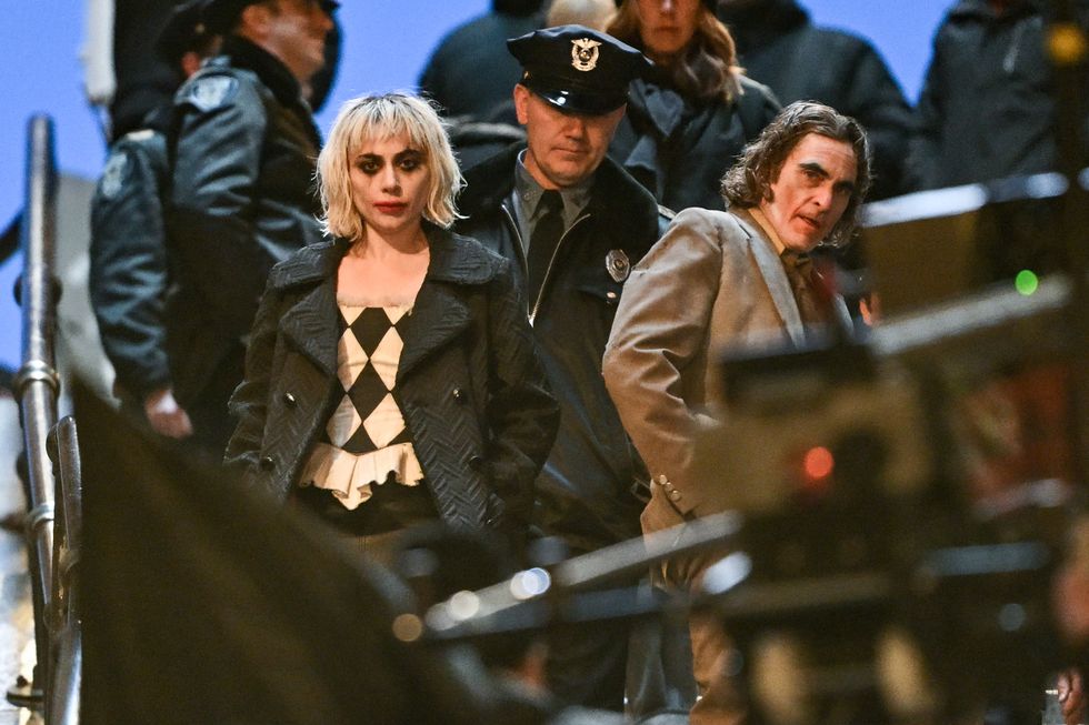 lady gaga and joaquin phoenix star at the camera while in character for the joker sequel, she wears a gray peacoat with a black and white diamond patterned shirt, he wears a brown suit, both have face paint on and a fake police officer stands behind them