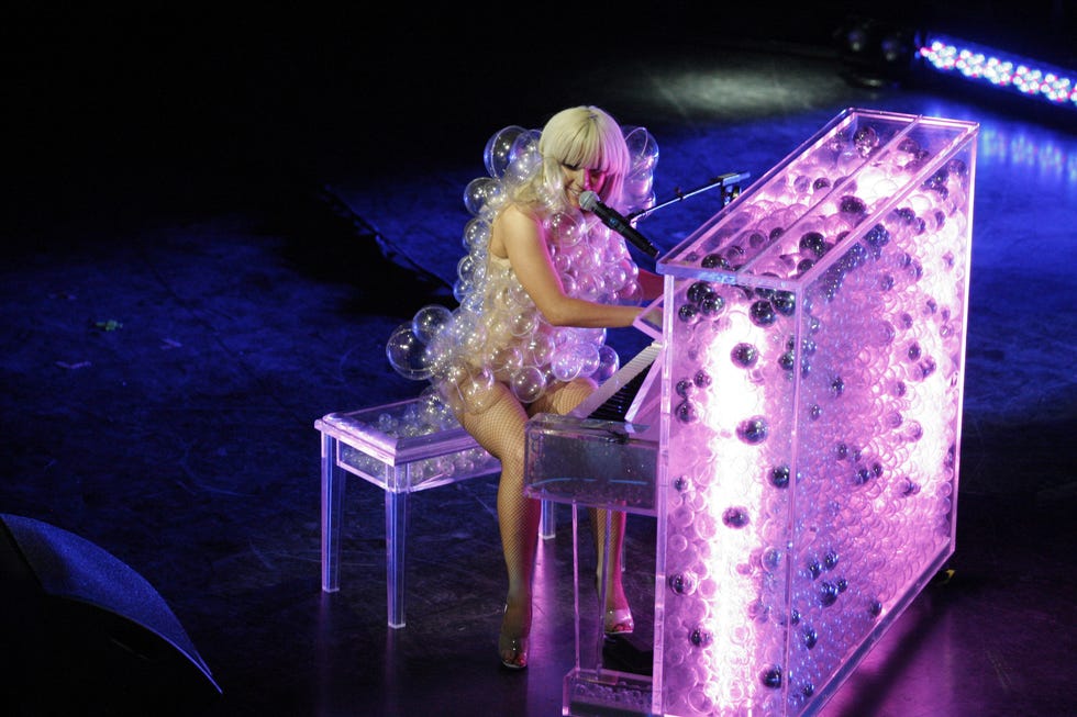 lady gaga plays piano and sings into a microphone on a stand while wearing a bubble suit