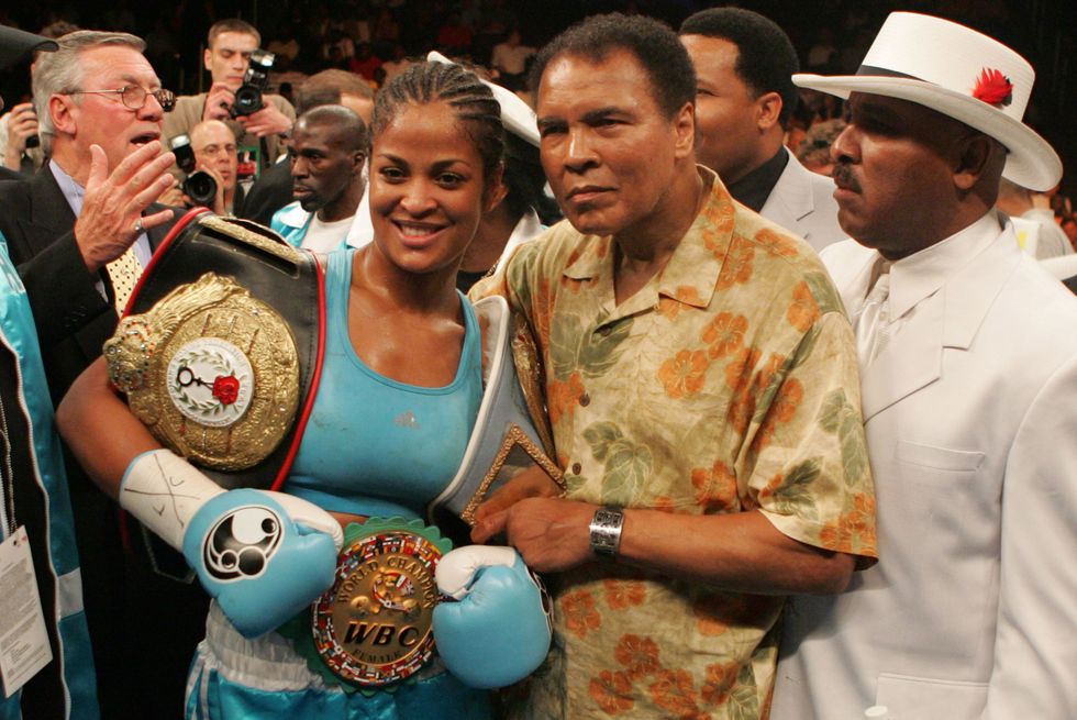 laila ali and muhammad ali pose for a photo after laila won the ﻿super middleweight title, which is represented by the large belt on her left shoulder, laila wears her boxing gloves and holds a medal in front of her while muhammad embraces her, behind them is a crowd of people