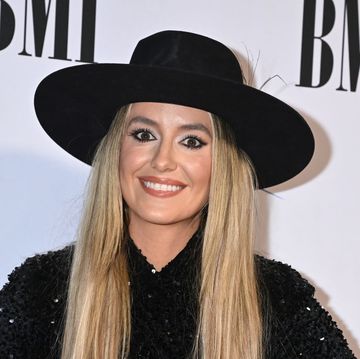 lainey wilson smiles at camera, she wears a black wide brimmed hat with a black sequin top