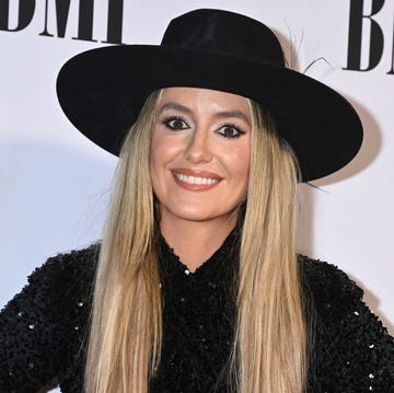 lainey wilson smiles at camera, she wears a black wide brimmed hat with a black sequin top