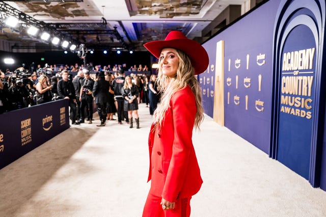 lainey wilson smiles at the camera while looking over one shoulder, she stands on a tan carpet in an all red outfit as photographers and other people stand in the background