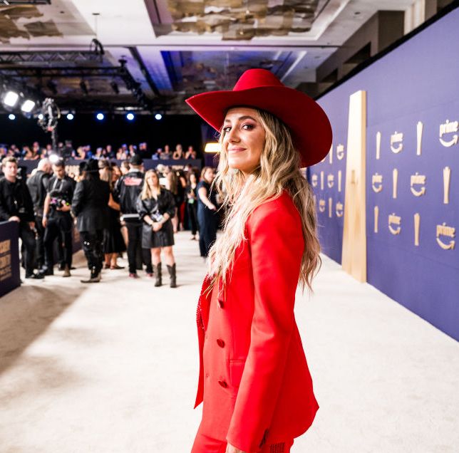 lainey wilson smiles at the camera while looking over one shoulder, she stands on a tan carpet in an all red outfit as photographers and other people stand in the background