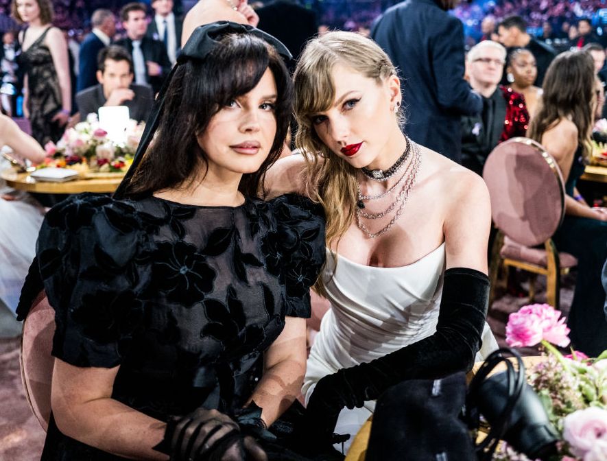 lana del rey and taylor swift pose for a photo while sitting at a table in a crowded room, lana wears all black and taylor wears a white dress with black gloves