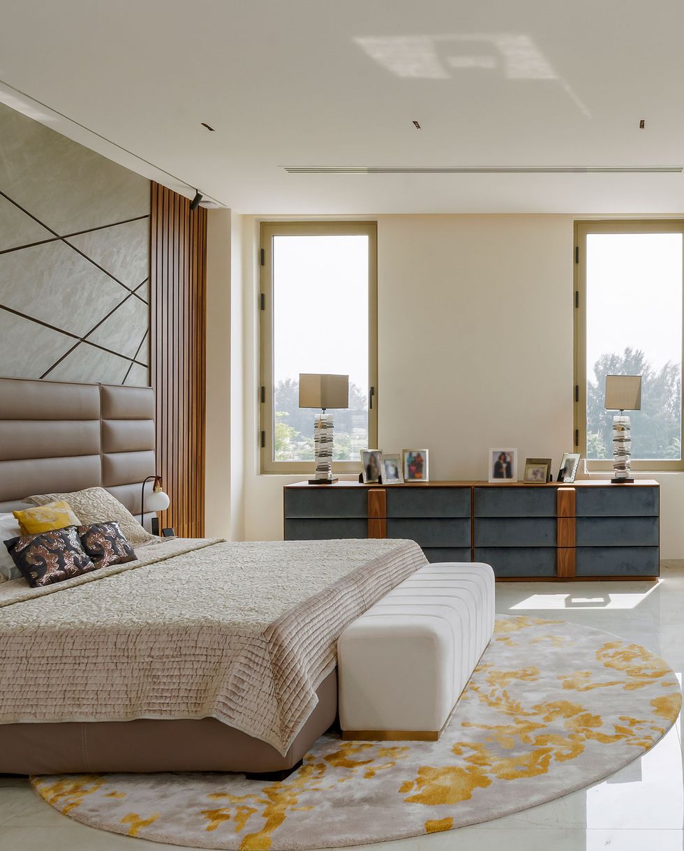 bedroom suite with king bed with brown vertically tufted leather headboard and a circular yellowish white rug underneath on polished marble floors and a long console opposite and a tv mounted above and two nice size windows at the back with anther console piece in between them