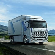 white hyundai xcient fuel cell electric truck driving on the asphalt road next to the green field in rural landscape at sunset