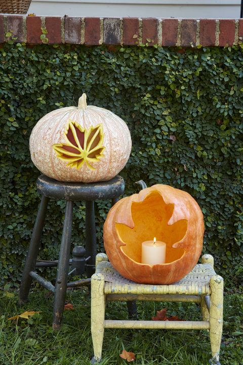 two pumpkins, each with large fall leaf design carved on front, displayed on rustic stools against vine covered wall