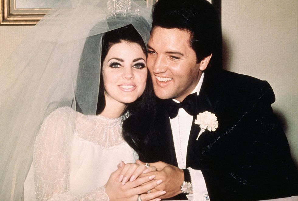 priscilla presley and elvis presley smile and hold hands, she wears a white lace wedding dress, white veil, and silver crown, he wears a black tuxedo and matching bow tie with a white boutonniere on his lapel