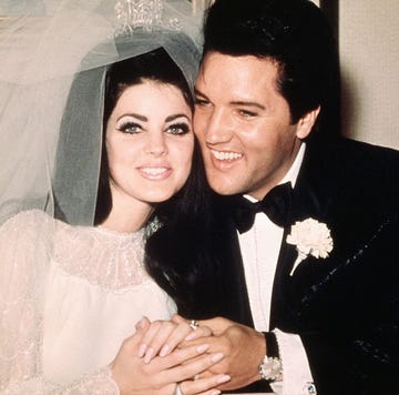 priscilla and elvis presley sit smiling and holding hands, she wears a wedding dress, veil and tiara, he wears a black suit with a bow tie and a carnation on his lapel