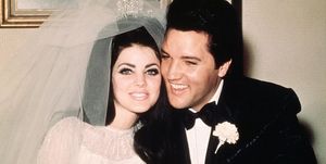 priscilla and elvis presley sit smiling and holding hands, she wears a wedding dress, veil and tiara, he wears a black suit with a bow tie and a carnation on his lapel