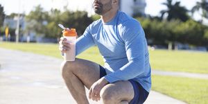 a latinx millennial male with a goatee, holds a travel mug with a smoothie, while wearing athletic clothing in an outdoor park