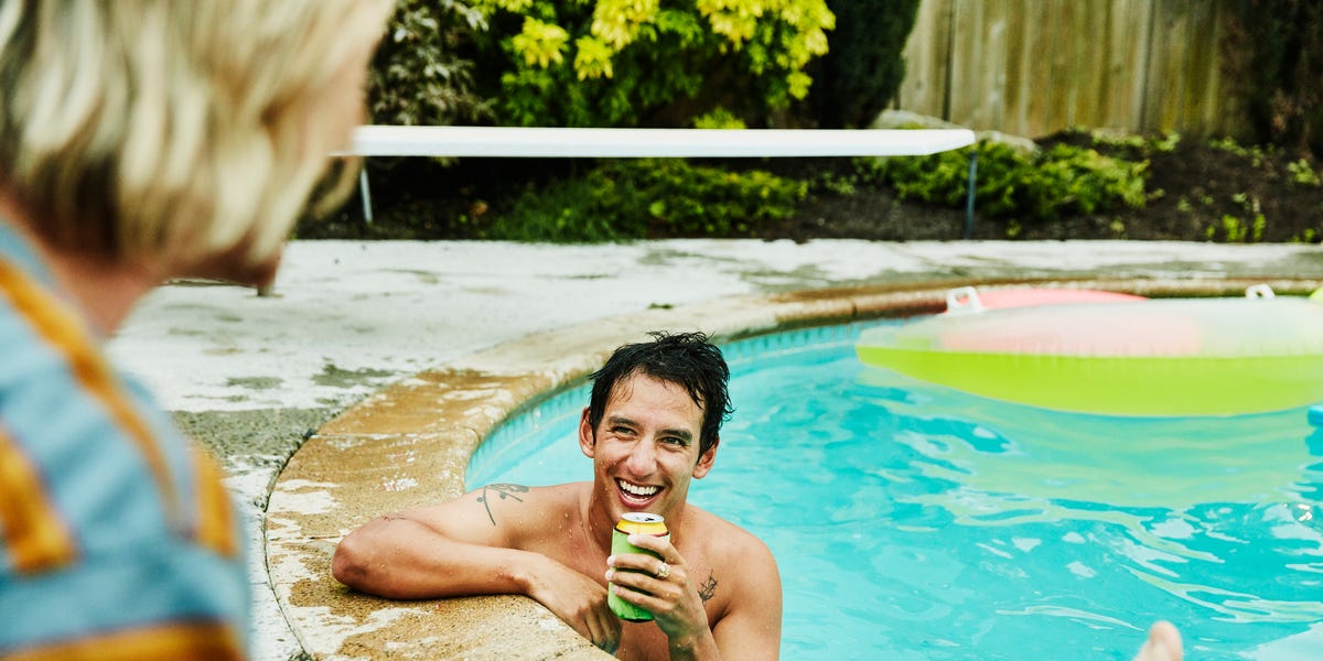 laughing man with drink in backyard pool in discussion with friend on summer evening