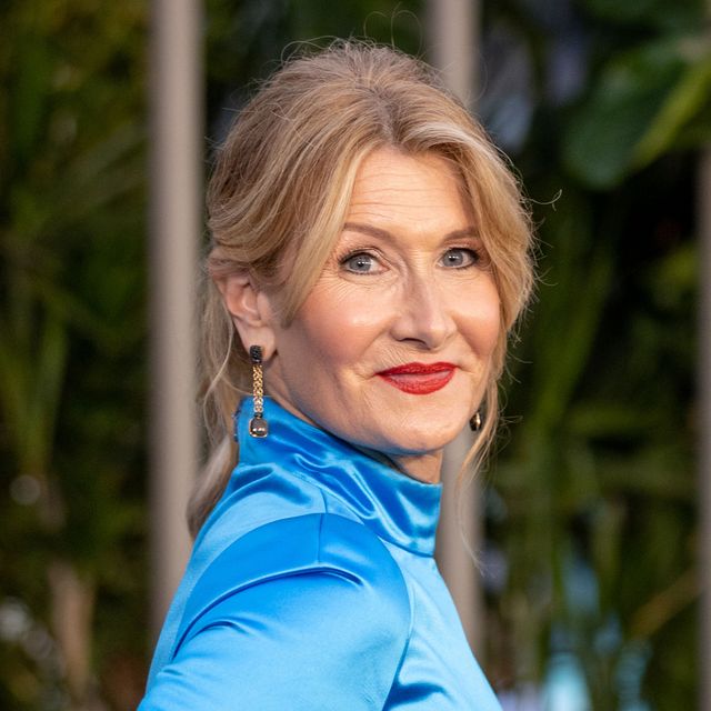 laura dern smiles at the camera as she looks over her shoulder, she wears a blue top with dangling earrings