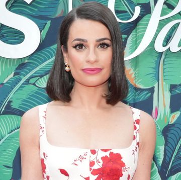 lea michele, a woman stands looking at the camera with a neutral facial expression, she has short brown hair to her shoulders and wears a white dress with red flowers on