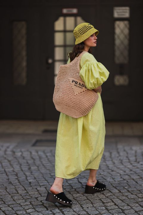 a woman wearing a yellow bubble maxi dress with black clogs, a prada straw bag, and yellow bucket hat on the street in berlin in a roundup of cute summer outfits 2023