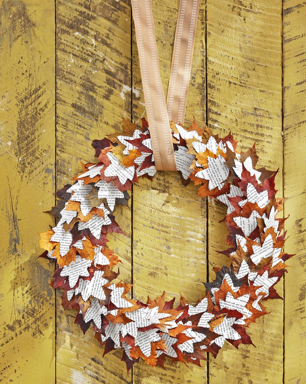 paper wreath crafted from fall leaves layered with leaf shapes cut from book pages, in front of yellow wall