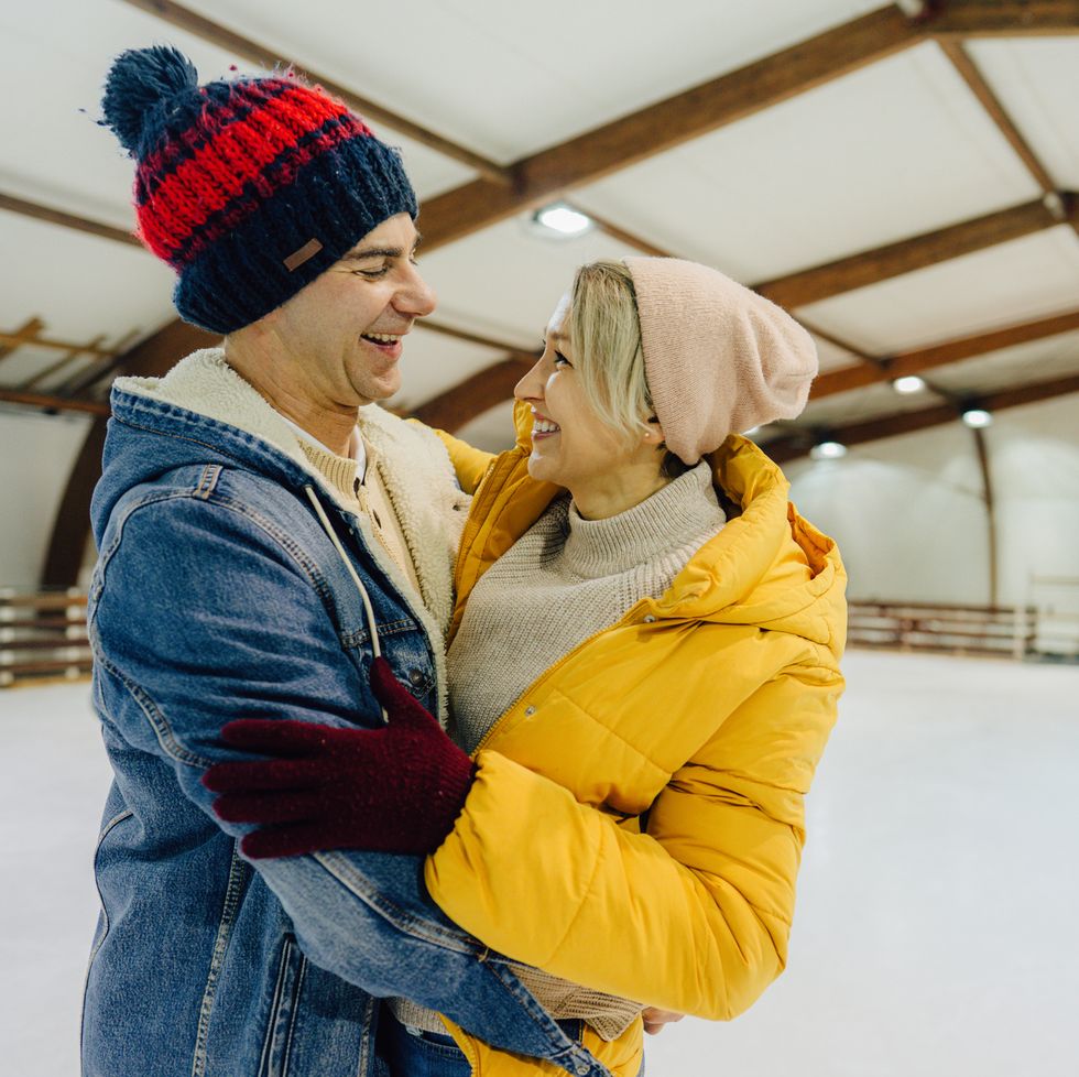 a white couple wearing winter attire hugging at an indoor skating rink