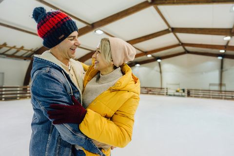 a white couple wearing winter attire hugging at an indoor skating rink