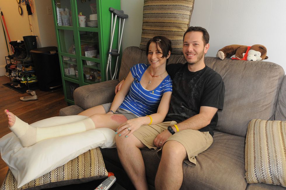 lee ann and nick yanni sitting on a brown couch in a living room and smiling, with lee ann's leg in a cast resting atop pillows