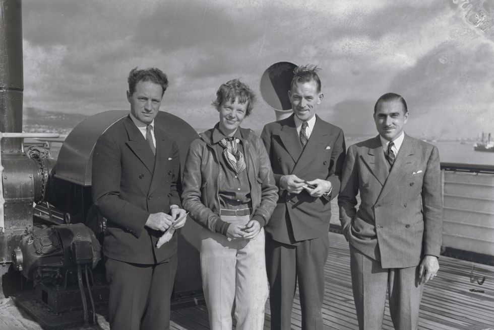 harry manning, amelia earhart, fred noonan, and paul mantz smile and stand on a ship deck, the men wear suits, earhart wears a leather jacket, collared shirt, scarf, and slacks