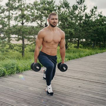 motivated bearded man with naked torso makes squat with heavy barbells, does physical exercises outdoor, enjoys workout, poses near green trees, doing weightlifting healthy lifestyle concept