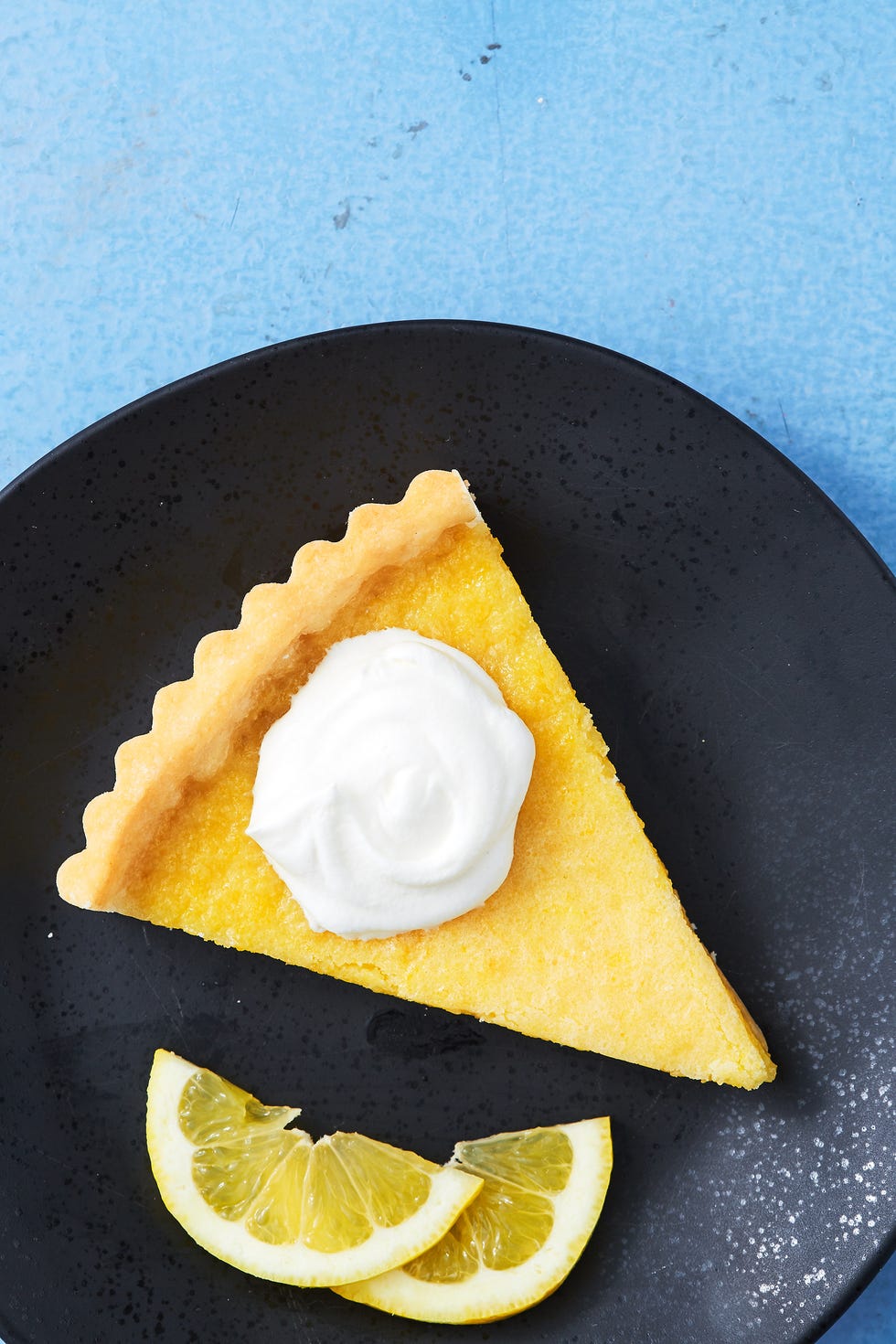 slice of lemon tart topped with whipped creme on a black plate with two lemon slices