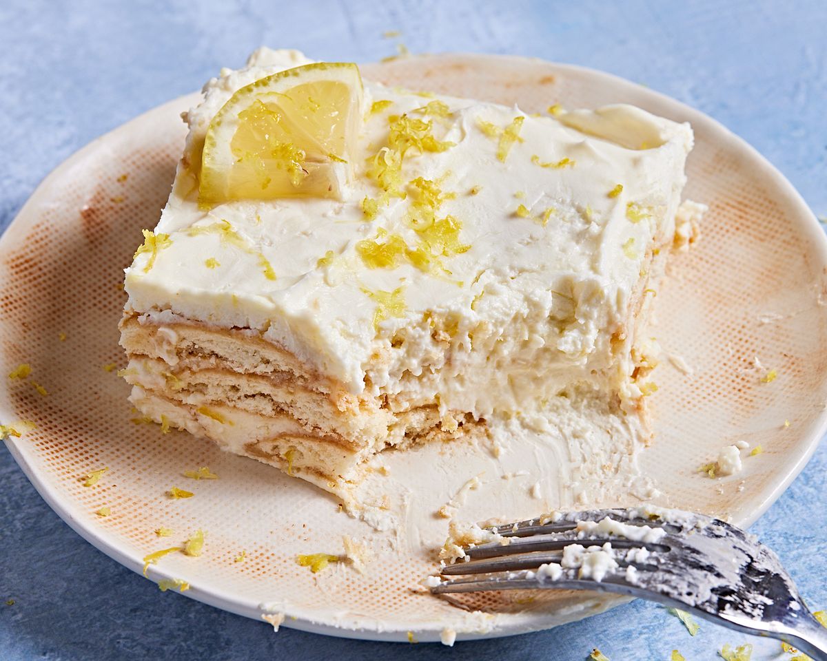 square slice of lemon icebox cake with a bite out on an off white plate on a blue background