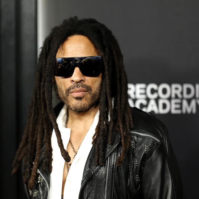 lenny kravitz looks at the camera, he wears dark tinted sunglasses, a black leather jacket and a white shirt with necklaces, he stands in front of a black background