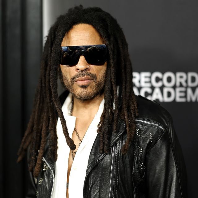 lenny kravitz looks at the camera, he wears dark tinted sunglasses, a black leather jacket and a white shirt with necklaces, he stands in front of a black background