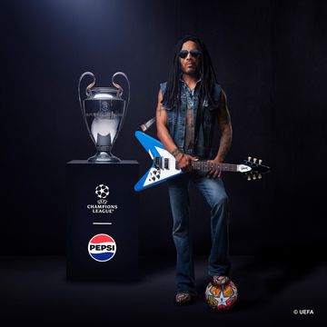 lenny kravitz holding a guitar with a football under his feet, next to him is the silver champions league trophy