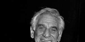 leonard bernstein smiles at the camera, he wears a wool coat and three piece suit with a white collared shirt and tie, he holds a pair of glasses in his right hand