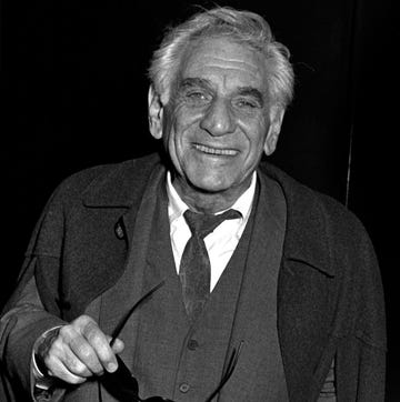 leonard bernstein smiles at the camera, he wears a wool coat and three piece suit with a white collared shirt and tie, he holds a pair of glasses in his right hand