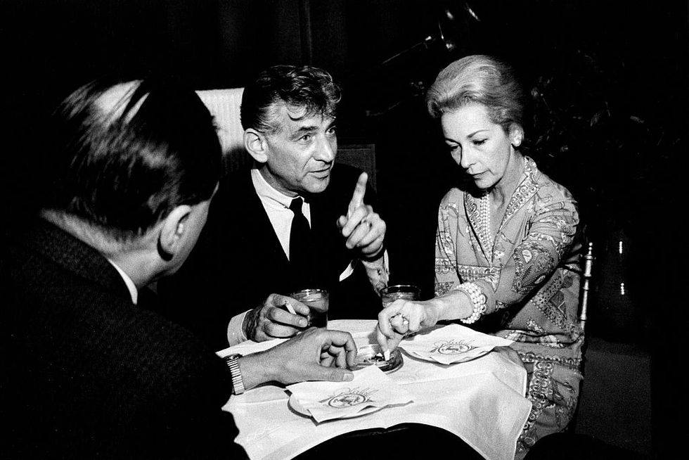 a black and white photo of leonard bernstein and felicia montealegre sitting at a table with another man, with leonard talking to someone off screen and pointing up, and felicia putting out a cigarette in an ashtray