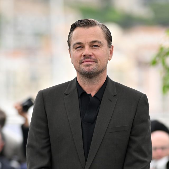 leonardo dicaprio stands and looks at the camera with a slight smile, he wears a gray suit jacket and black collared shirt, the outdoor background is blurred behind him